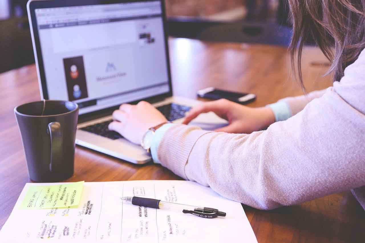 Eine Frau schreibt auf ihrem Laptop, daneben steht eine Tasse Kaffee.